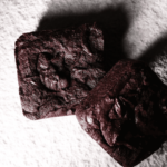 Two rich, fudgy vegan brownies on a textured beige surface with dramatic shadows.