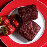 Two rich plant-based chocolate brownies served with fresh strawberries drizzled in chocolate, on a red and white plate